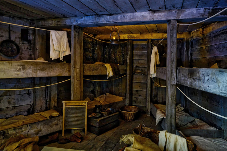Dimly lit, rustic cabin with wooden bunk beds, hanging laundry, a lantern, a washboard, and storage baskets.
