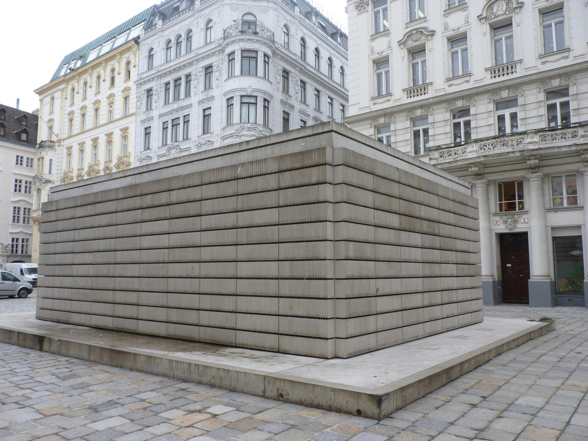 En stor rektangulär betongkonstruktion i en kullerstensplaza, omgiven av gamla byggnader i europeisk stil.