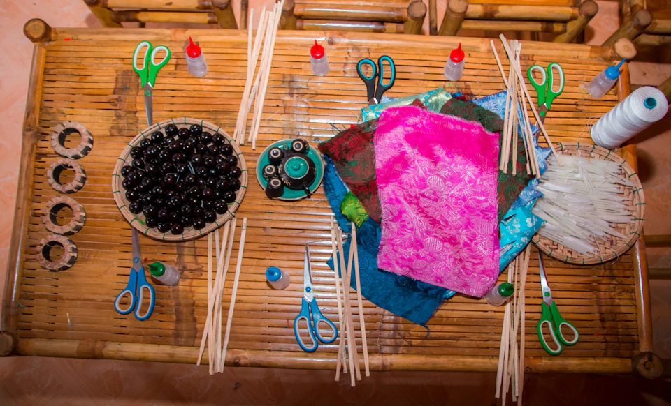 A wooden table with craft supplies, including fabric, scissors, sticks, glue bottles, and a bowl of dark beads.