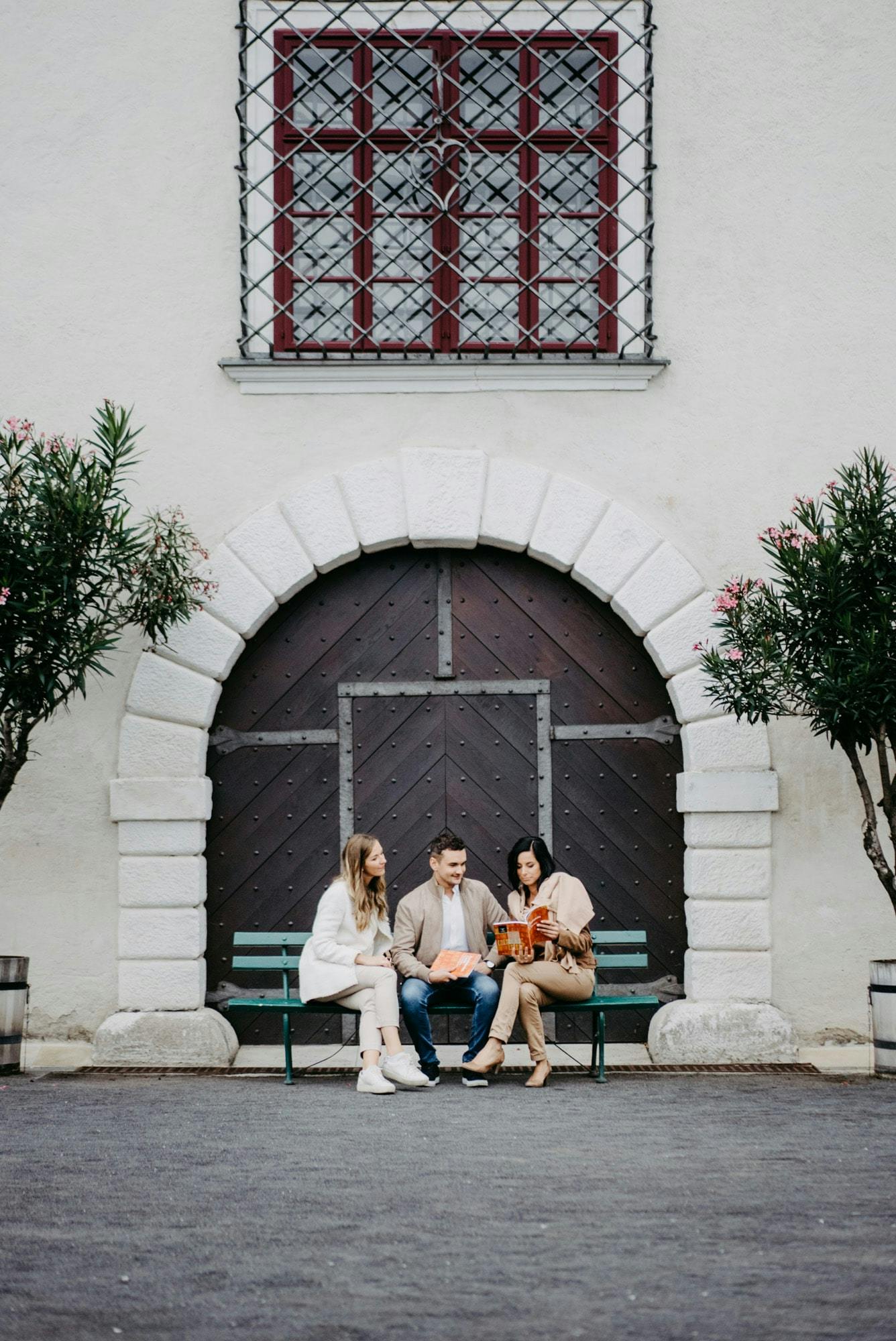 돌담에 세워진 아치형 나무 문 앞 벤치에는 나무 두 그루 옆에 세 사람이 앉아 있습니다.