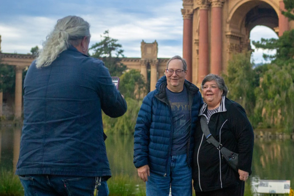 Two people posing for a photo near a scenic landmark with tall columns and greenery in the background.