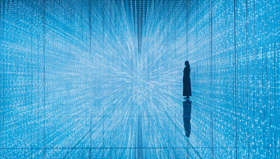 A person stands amidst a mesmerizing, illuminated room with blue lights creating a tunnel-like, infinite pattern effect.