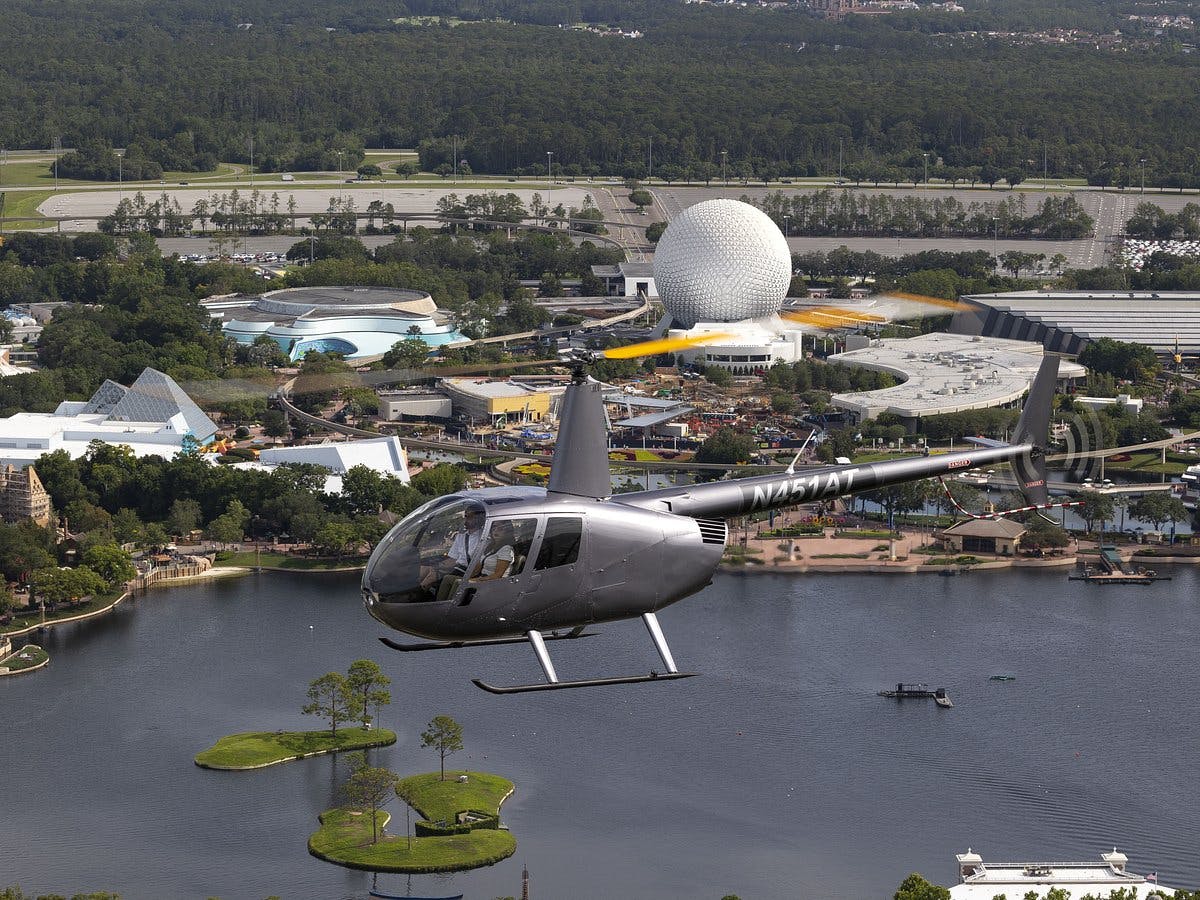 Vrtulník letící nad zábavním parkem s vodou, zelení a velkou geodetickou kopulí.