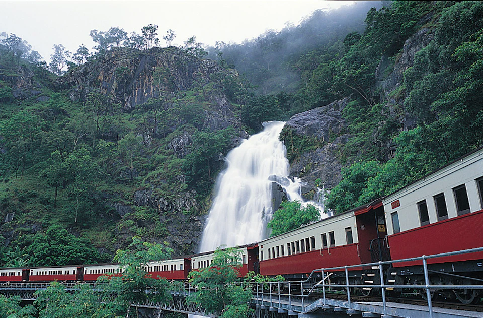 A red and cream train passes through a lush, mountainous area with a large waterfall in the background.
