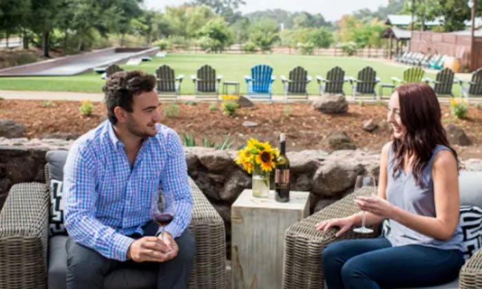 Muž sedí venku na proutěné židli a drží sklenici vína. V pozadí jsou židle Adirondack a svěží zahrada.