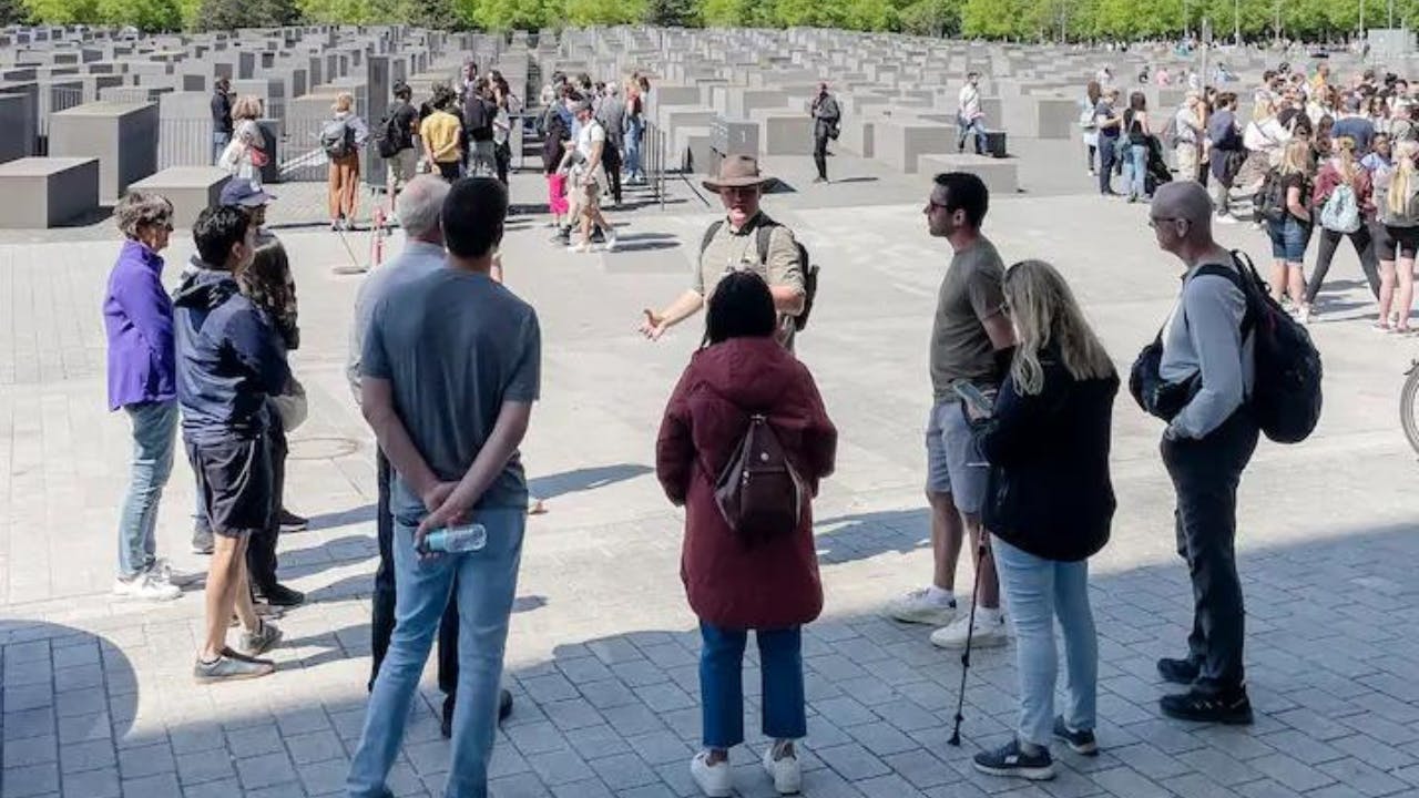 Skupina lidí poslouchá průvodce poblíž památníku s mnoha betonovými bloky. V pozadí je vidět více návštěvníků.