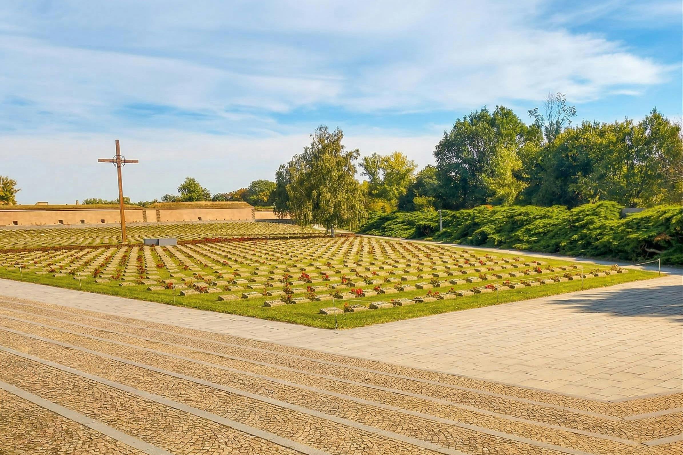 Ένα νεκροταφείο με σειρές τάφων, μερικοί με λουλούδια, ένα μεγάλο σταυρό και γύρω πράσινο κάτω από ένα γαλάζιο ουρανό.