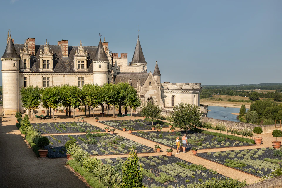 A historic castle with pointed towers overlooks a manicured garden with pathways and two people walking under a clear sky.
