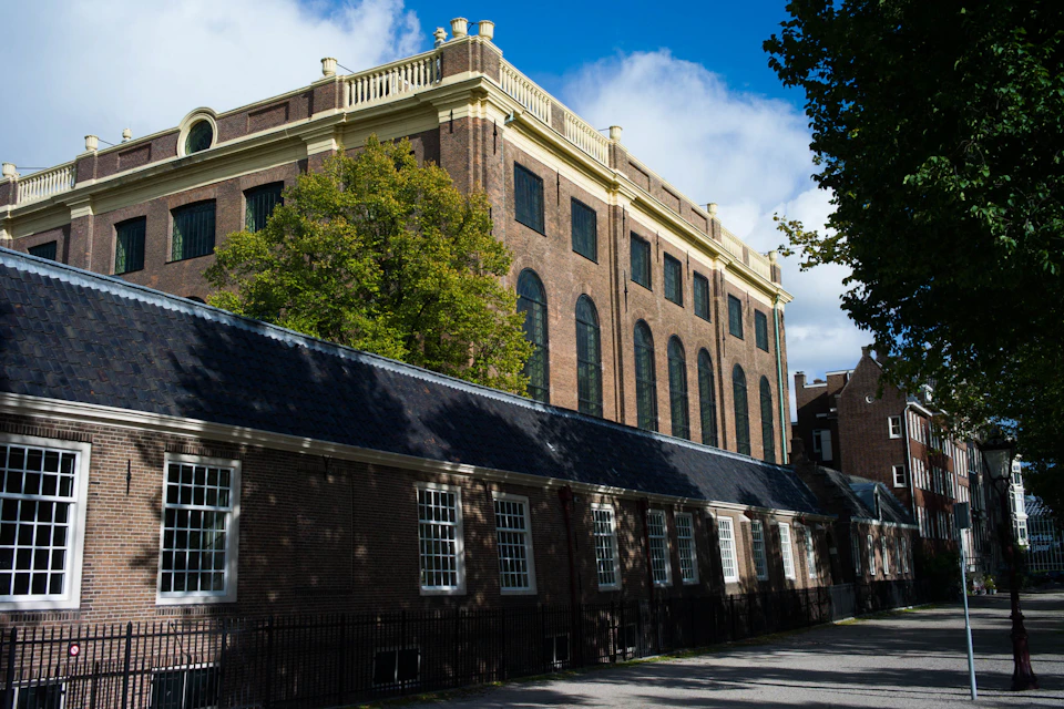 A historic brick building with large arched windows, adjacent to a lower structure, under a blue sky with scattered clouds.
