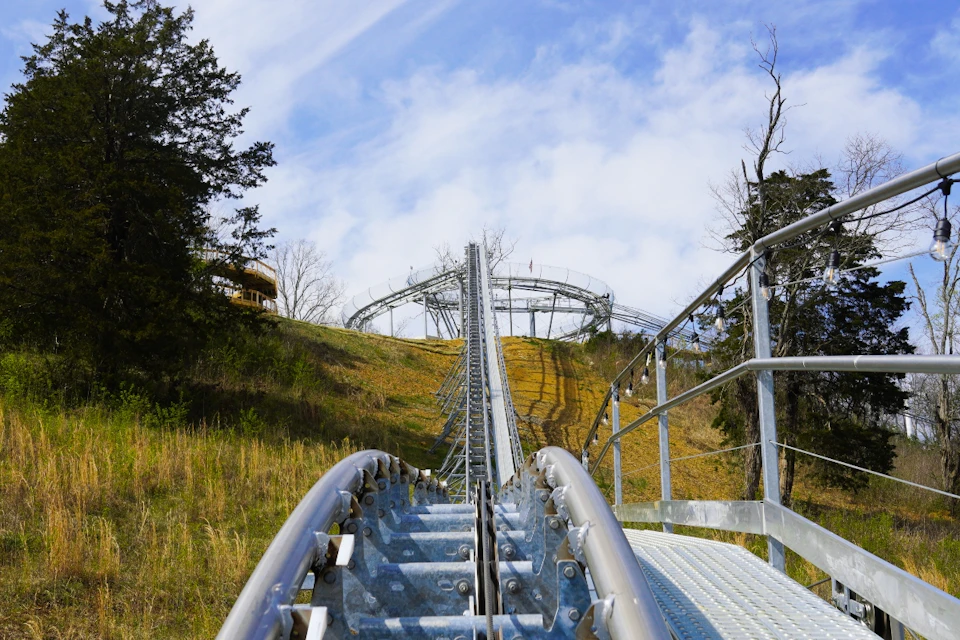 A metal roller coaster track ascending a hill, surrounded by trees and grassy land under a partly cloudy sky.