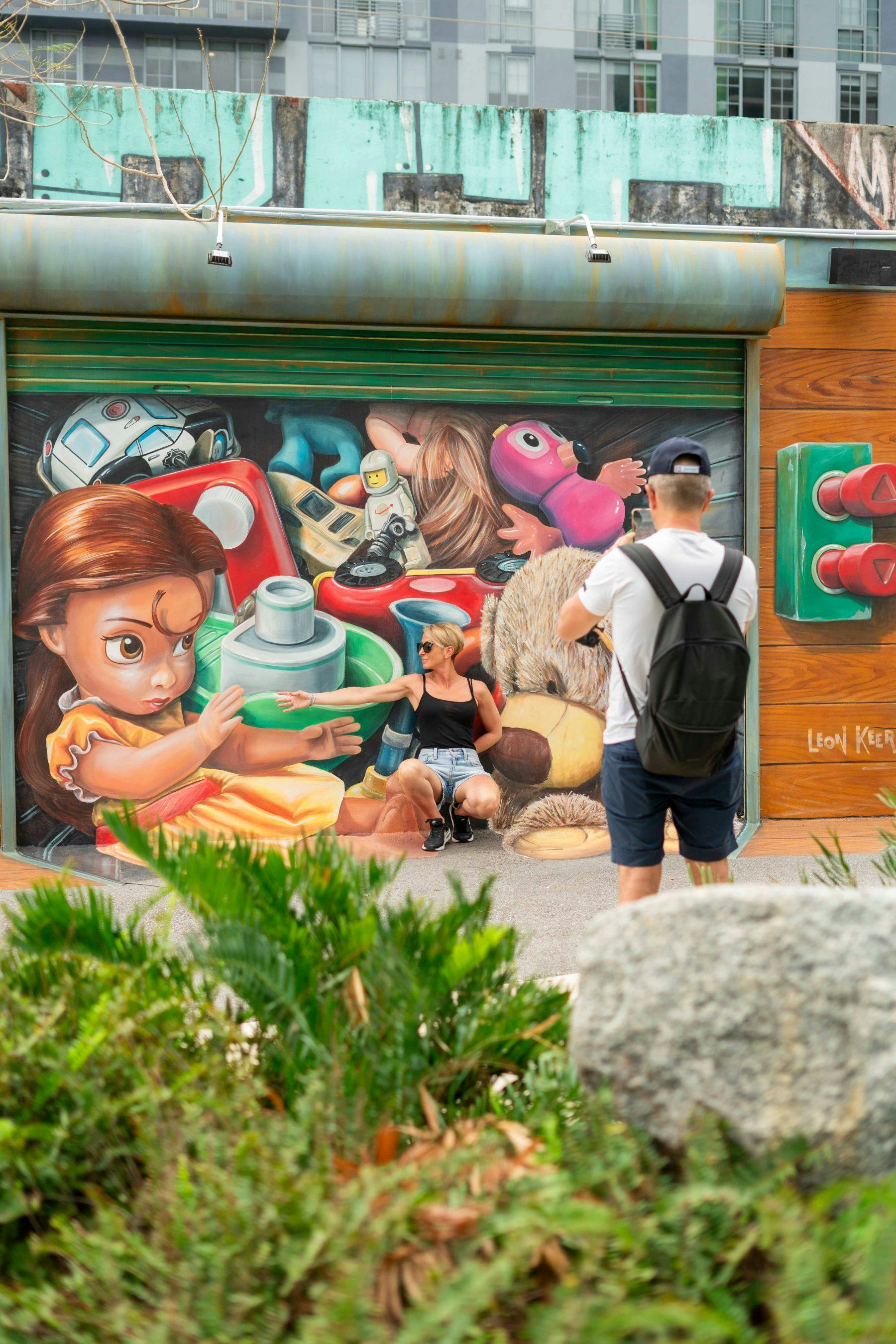 A person poses with a vibrant mural depicting toys, including a large doll, while another person photographs them.