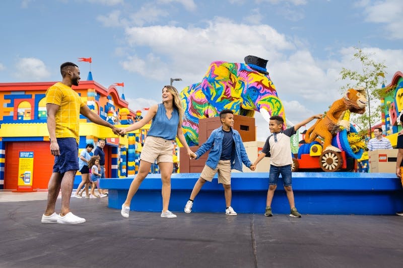 People enjoying an outdoor scene with colorful Lego structures, including an elephant and a lion on a float, against a blue sky.