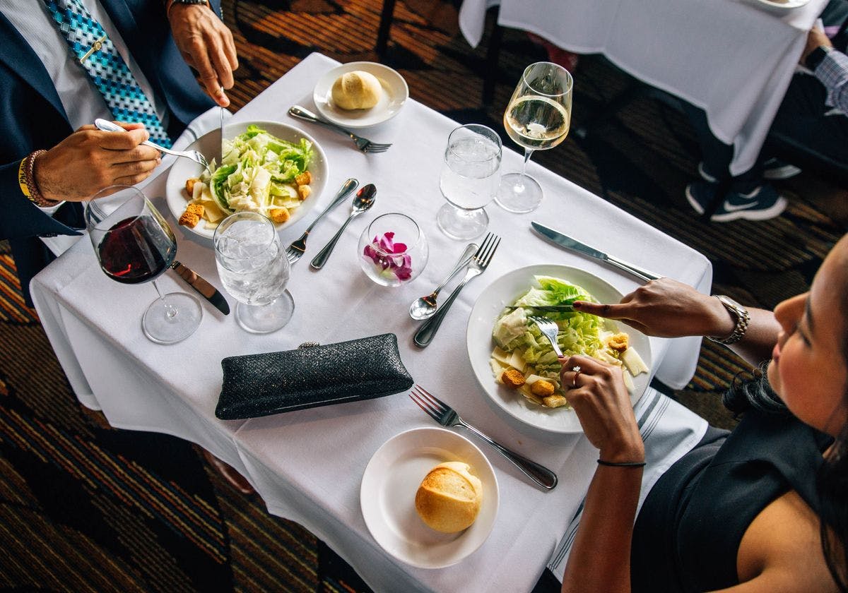Two people dining at a table with salads, bread rolls, wine glasses, and water glasses on a white tablecloth.