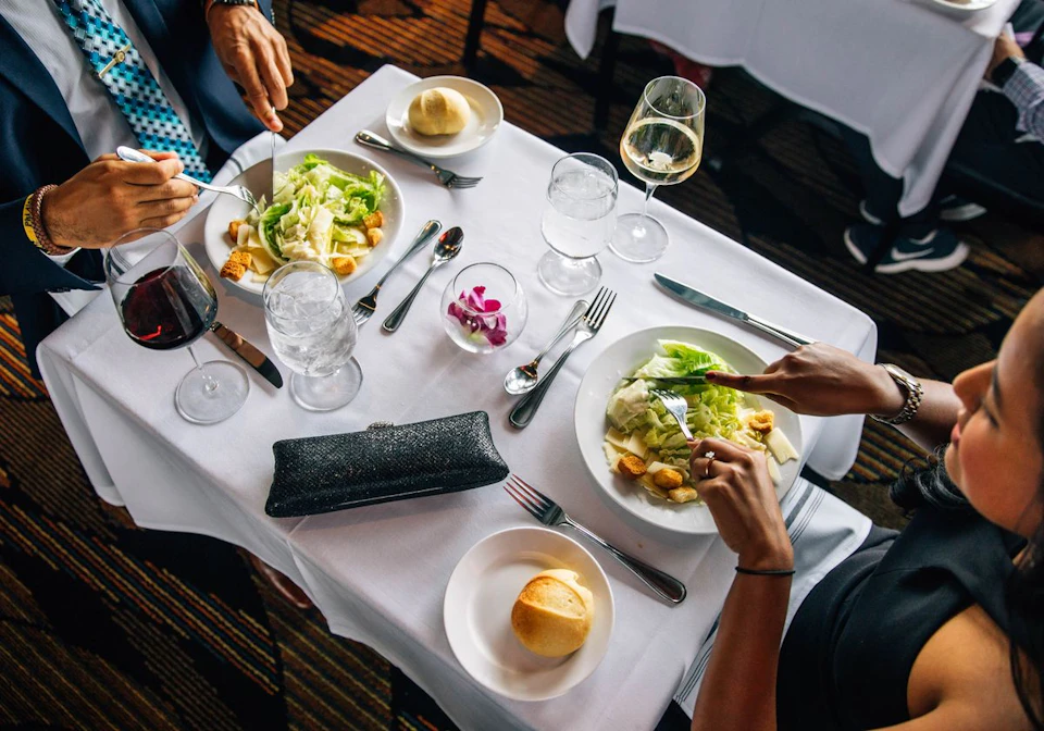 Two people dining at a table with salads, bread rolls, wine glasses, and water glasses on a white tablecloth.