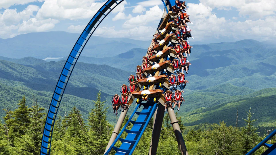 A rollercoaster with riders ascending a loop, surrounded by a forested mountain landscape under a blue sky with clouds.
