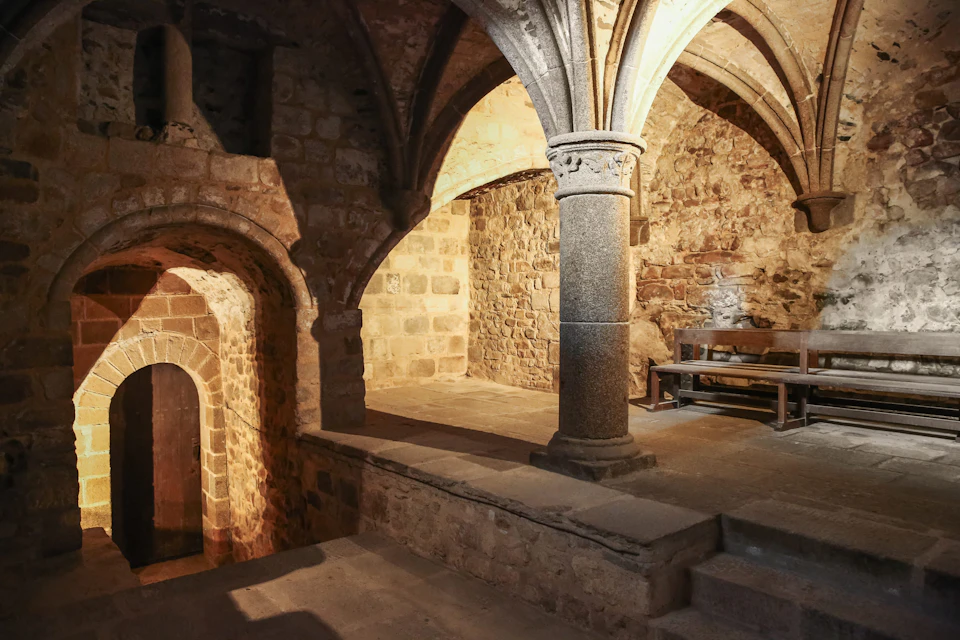 Ancient stone room with arched ceilings, pillars, and a wooden bench. Warm lighting highlights textured walls and doorways.