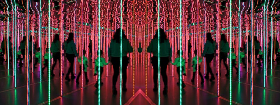 Persone che camminano in una stanza illuminata a specchio con luci verticali rosse e verdi che creano riflessi ripetuti.