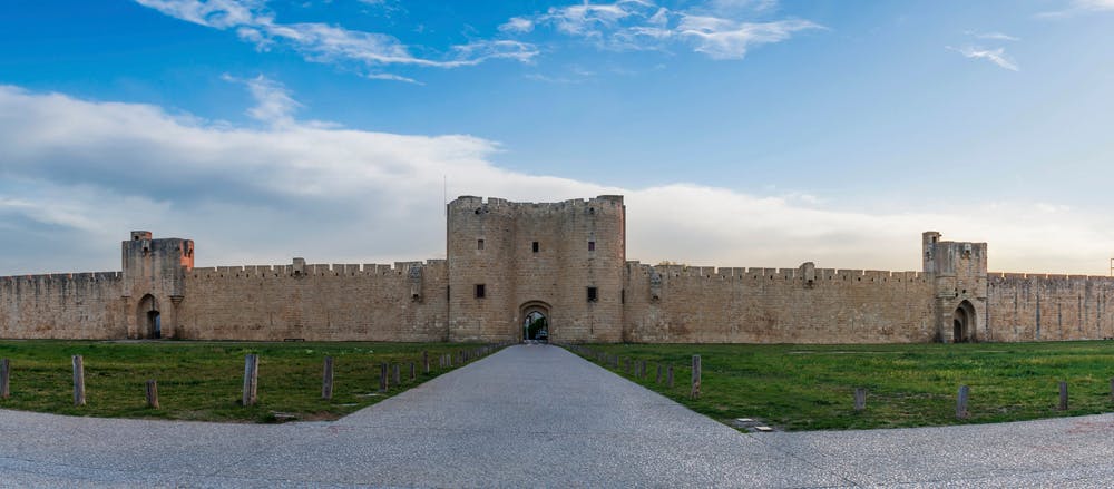 Stone fortress with a large central gate, flanked by high walls and towers, under a partly cloudy blue sky. A wide path leads to the gate.