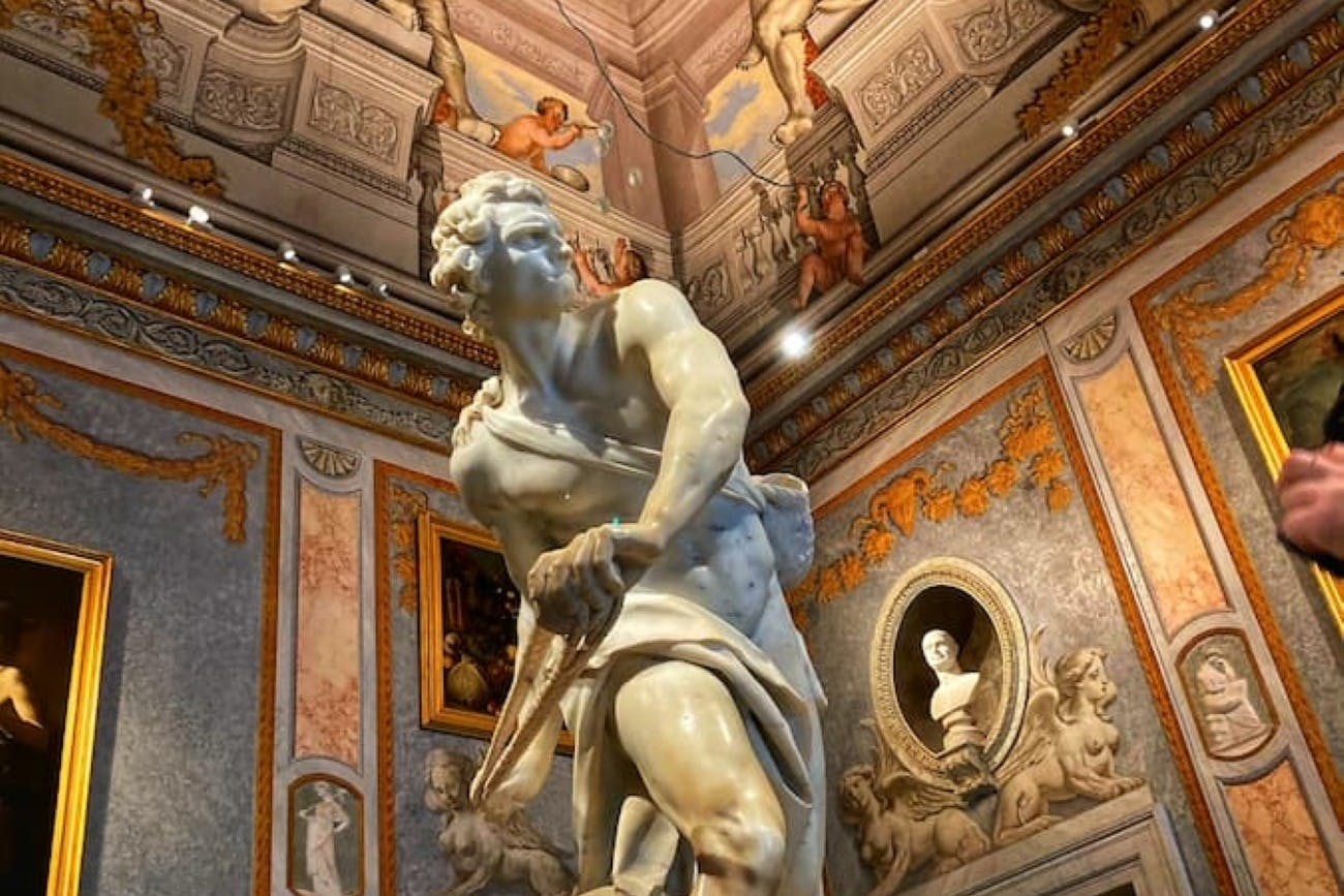 Una statua di marmo in una stanza riccamente decorata con pareti decorate, un soffitto dipinto e opere d'arte incorniciate, tra cui un busto in un'alcova.