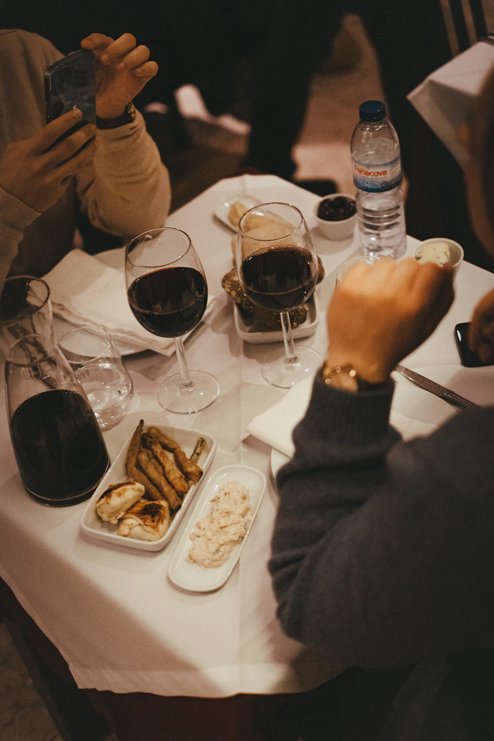 A table with plates of food, two glasses of red wine, a water bottle, and three people seated around it.