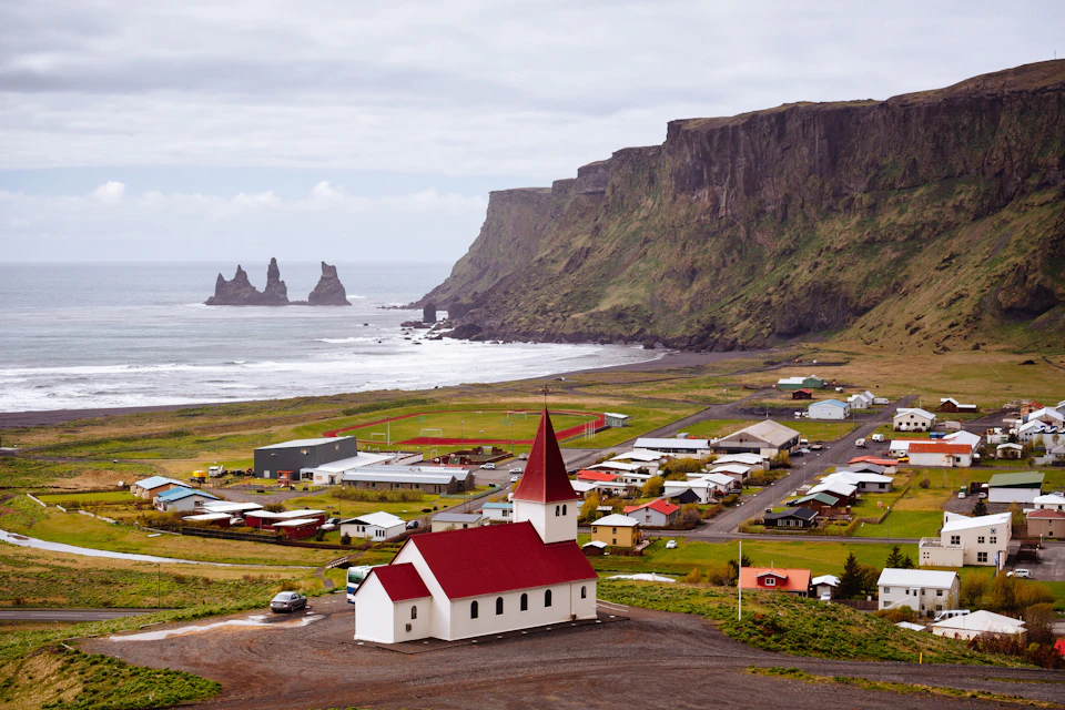 A small village with red-roofed church, fields, houses, and a coastal cliff with rock formations in the sea under a cloudy sky.