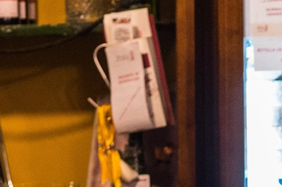 Four people talking inside a bar with hanging cured meats, red bows, and shelves of liquor and wine bottles.