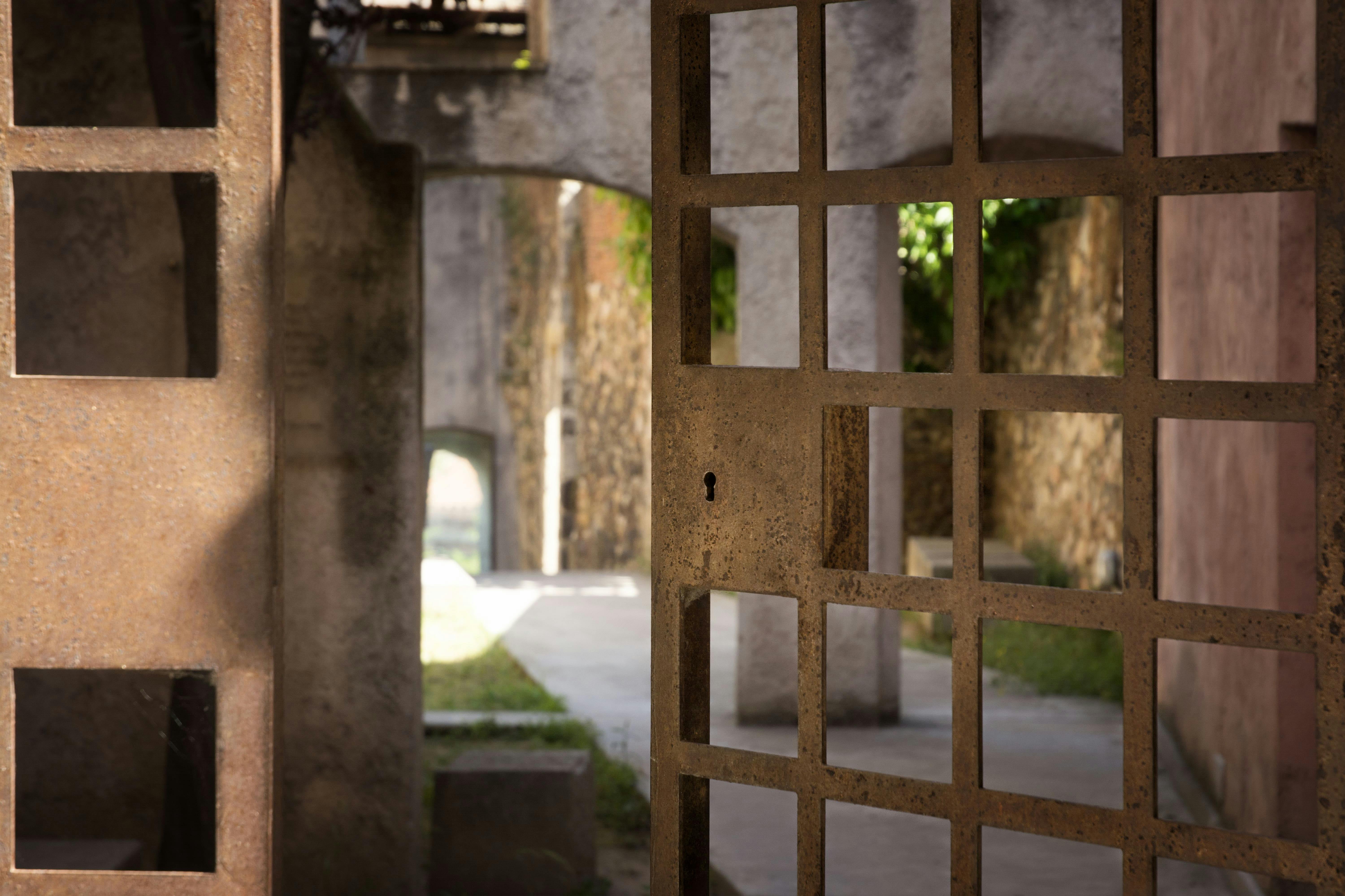 Rusty old gate with square cutouts, keyhole, and stone walls in the background. Sunlight illuminates the path beyond.