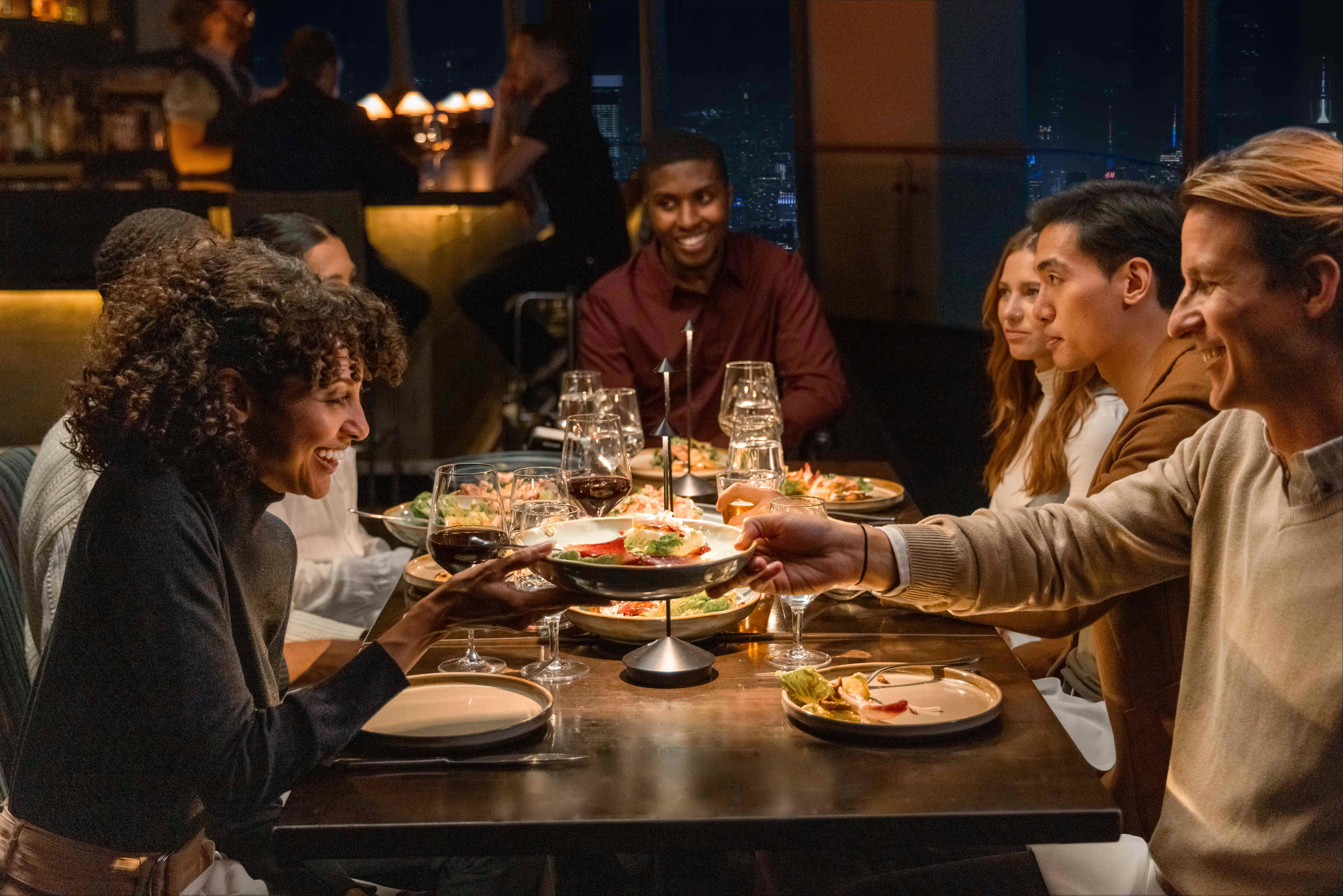 A group of people sharing food and smiling at a dinner table in a dimly lit restaurant.