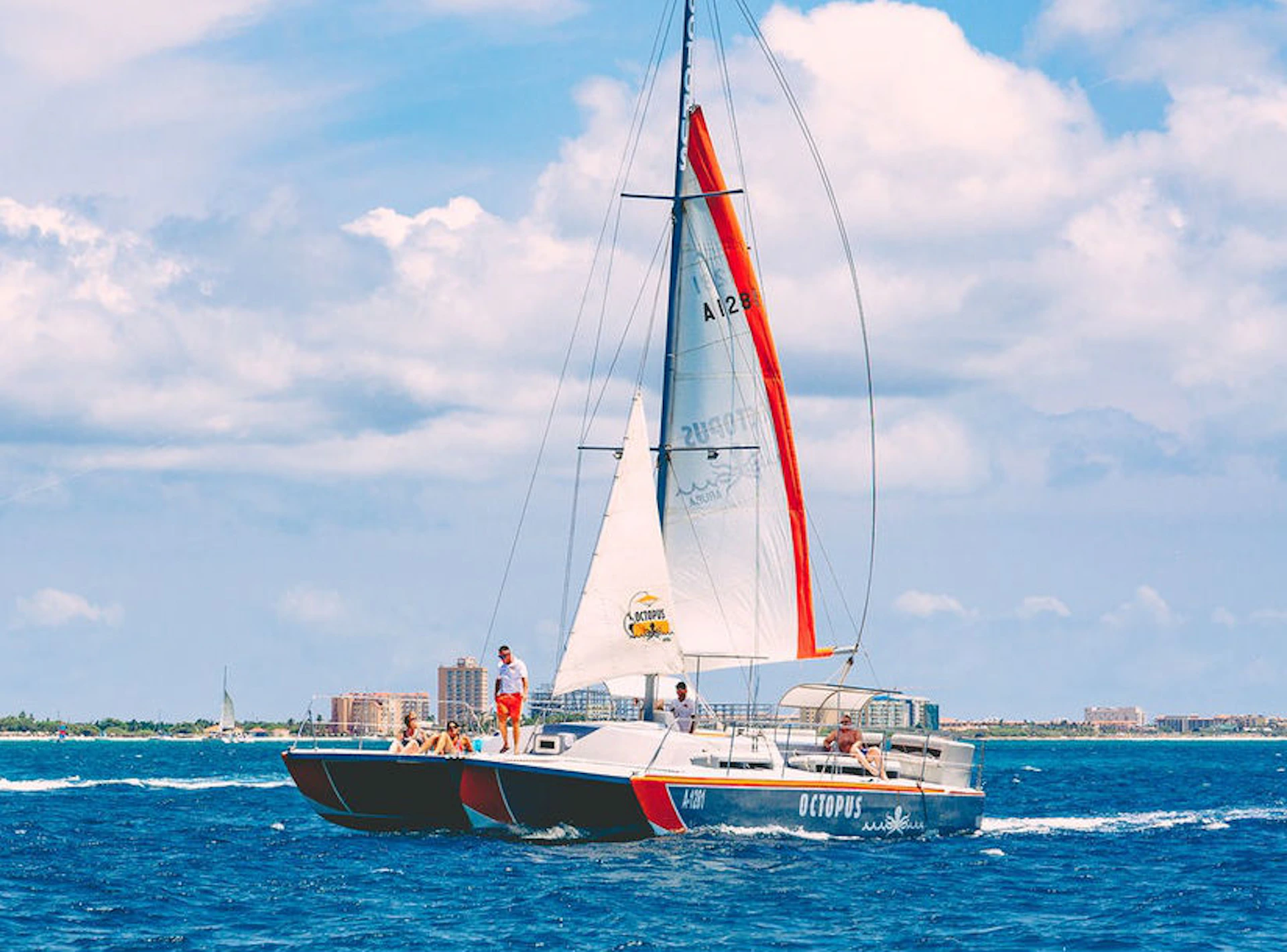 People relax on a catamaran named "OCTOPUS" sailing on a blue ocean under a partly cloudy sky, with distant buildings visible.