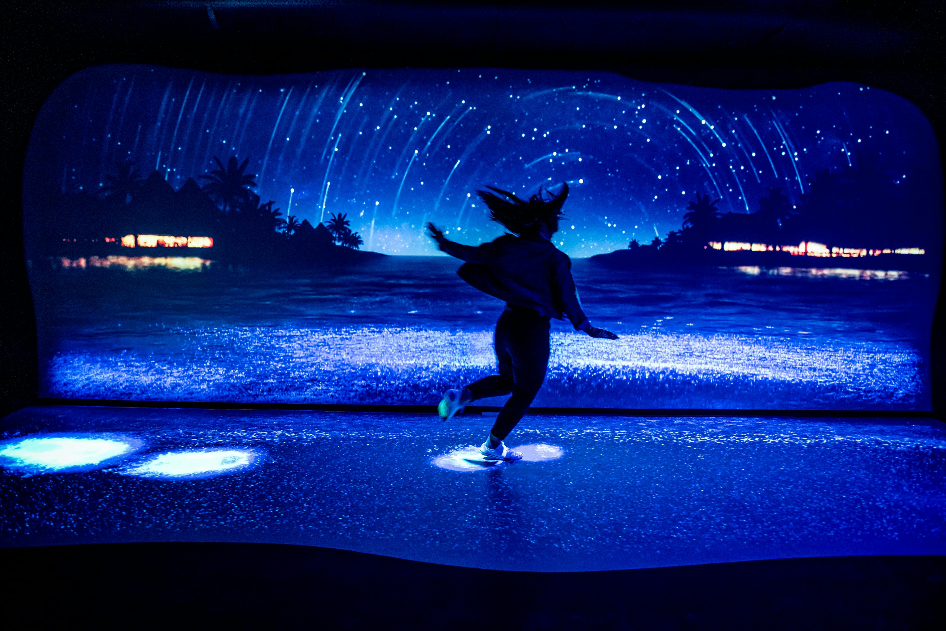 一个人在数字屏幕前跳舞，在海滩上展示星光熠熠的夜空，海滩上有轮廓鲜明的棕榈树和发光的灯光。