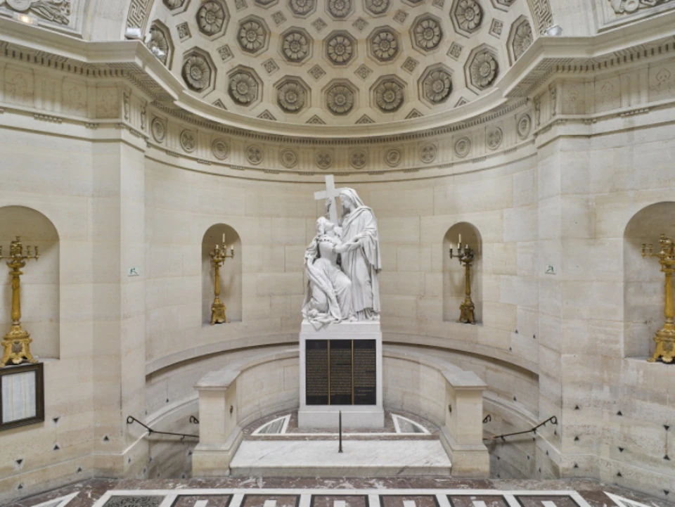An ornate interior with a central marble statue of a kneeling figure and a standing figure holding a cross, flanked by two gold candleholders.