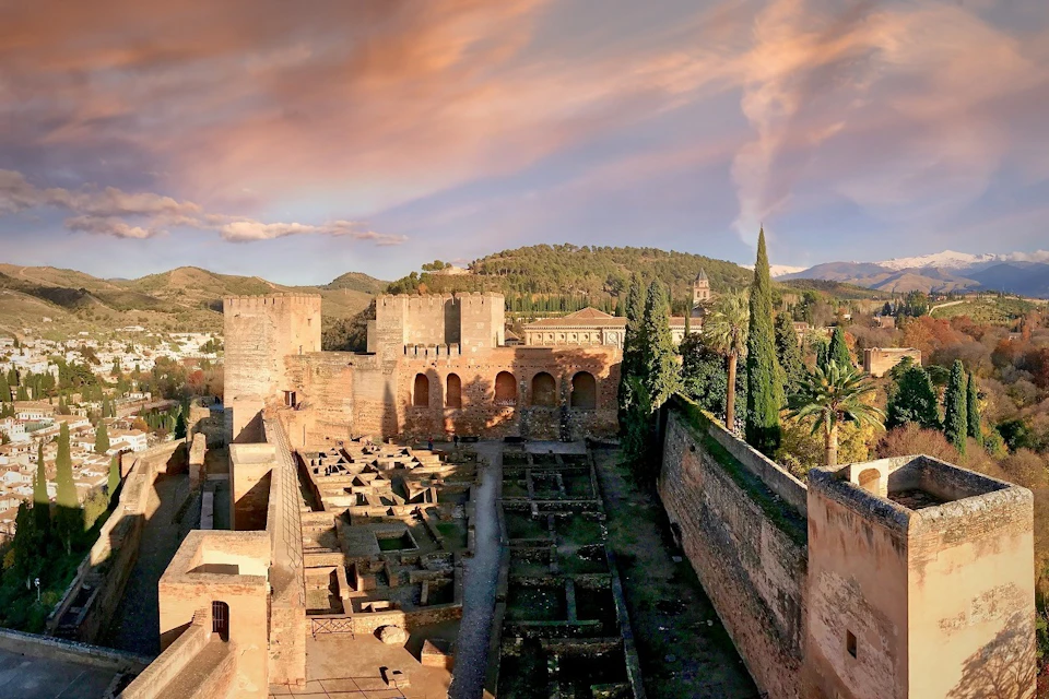 A historic castle complex featuring stone walls, towers, and courtyards, surrounded by lush greenery under a vibrant sky.