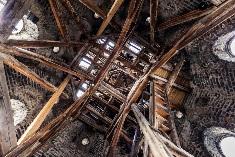 View of wooden beams and ladders forming a complex structure within a stone tower. Light enters through openings above.