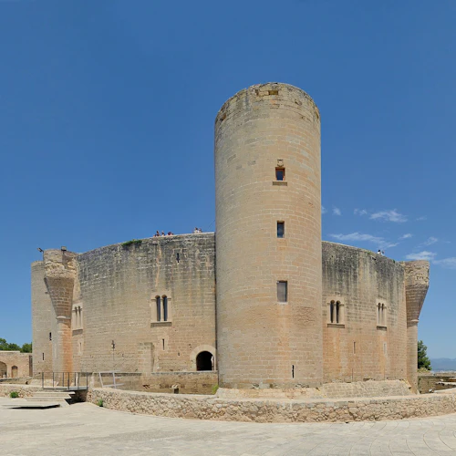 Ein steinernes Schloss mit einem hohen zylindrischen Turm unter strahlend blauem Himmel, umgeben von einem gepflasterten Innenhof und einigen Bäumen.