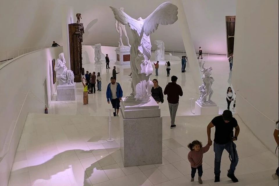 Visitors at a museum walk around marble sculptures in a bright, spacious hall with white walls and floors.