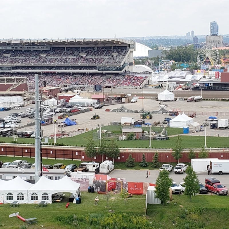 Overfyldt stadion med tilstødende markedsplads med forlystelser og telte. Parkeringsplads og flere bygninger synlige i forgrunden.