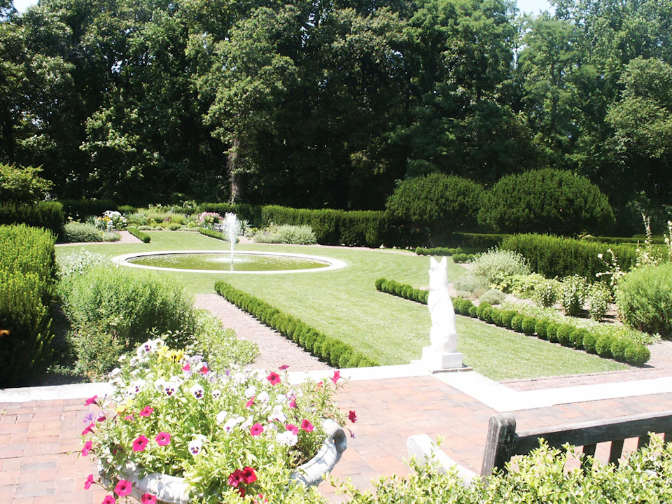 Manicured garden with blooming flowers, a central fountain, neatly trimmed hedges, and a stone animal statue under a sunny sky.