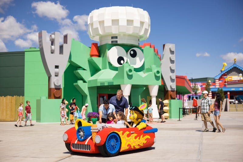 Adults and children interact with a Lego car. A large green dragon Lego structure stands in the background.