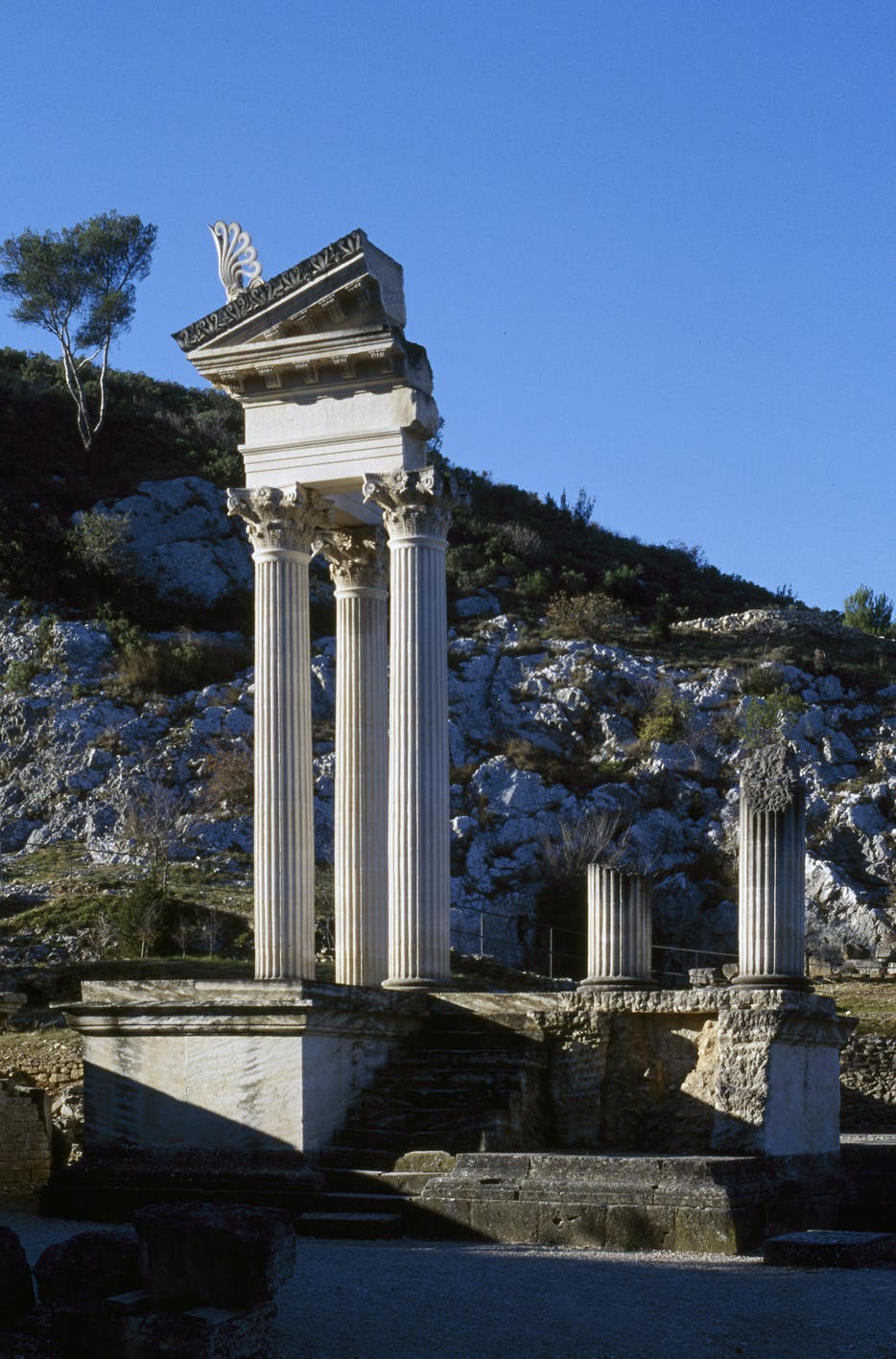 Antiche rovine con alte colonne bianche e una struttura parziale su un terreno roccioso sotto un cielo azzurro e limpido. Gli alberi sono sullo sfondo.