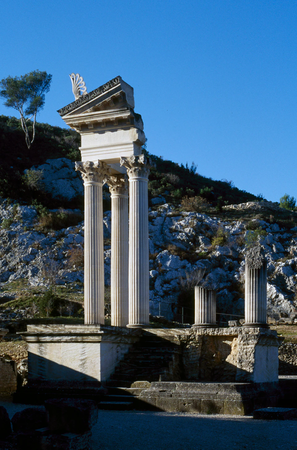 Ancient ruins with tall, white columns and a partial structure on rocky terrain under a clear blue sky. Trees are in the background.