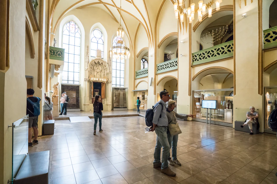 People explore a spacious, well-lit museum hall with arched windows, chandeliers, and historical artifacts on display.