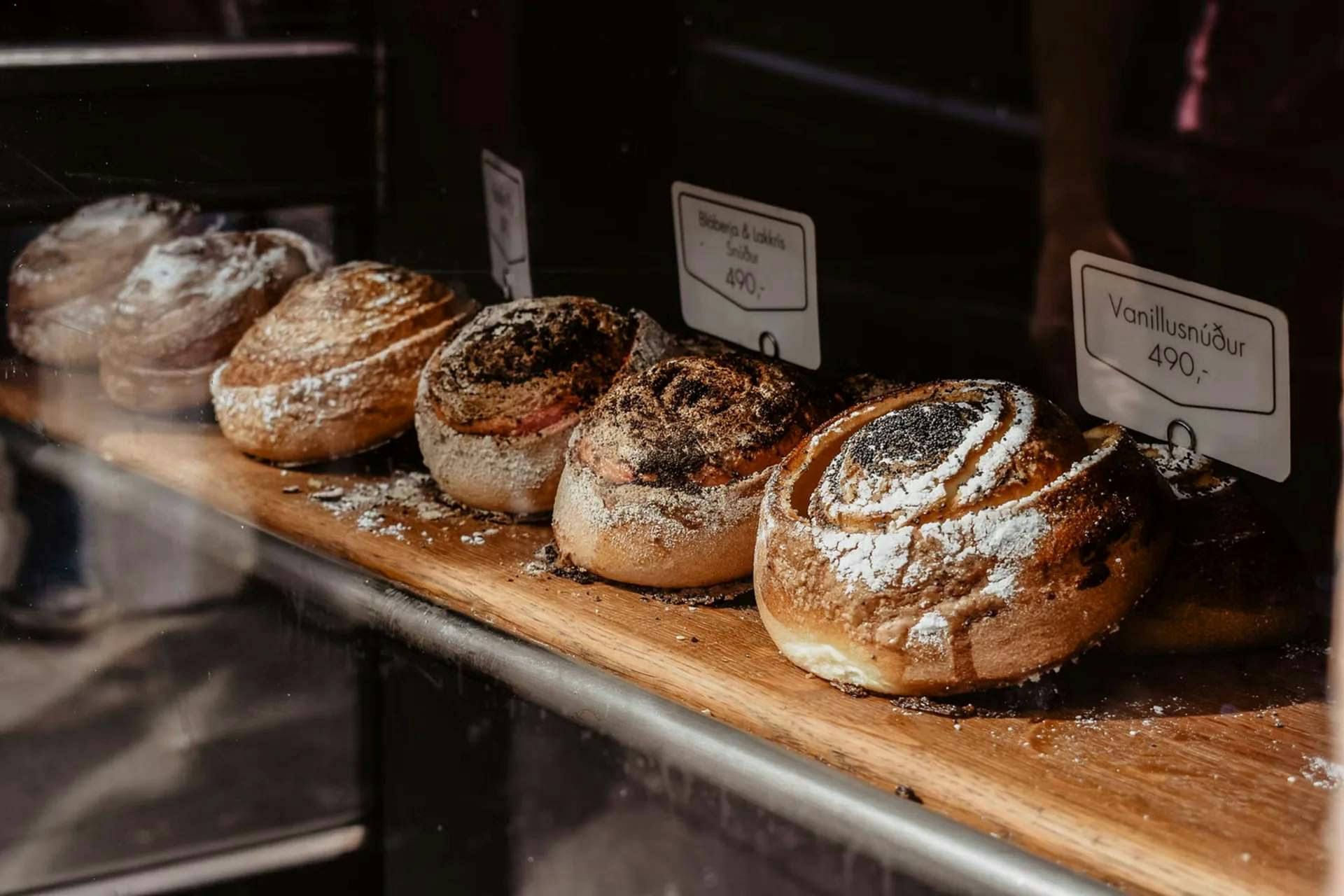 A row of pastries with powdered sugar and burnt tops displayed on a wooden board inside a showcase.