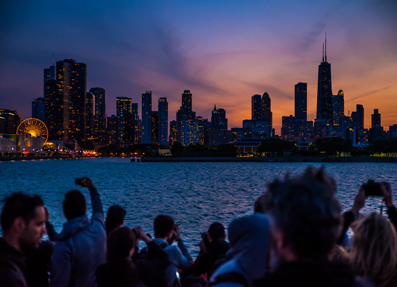 People on a boat taking photos of a city skyline at dusk, with a Ferris wheel and illuminated skyscrapers in the background.
