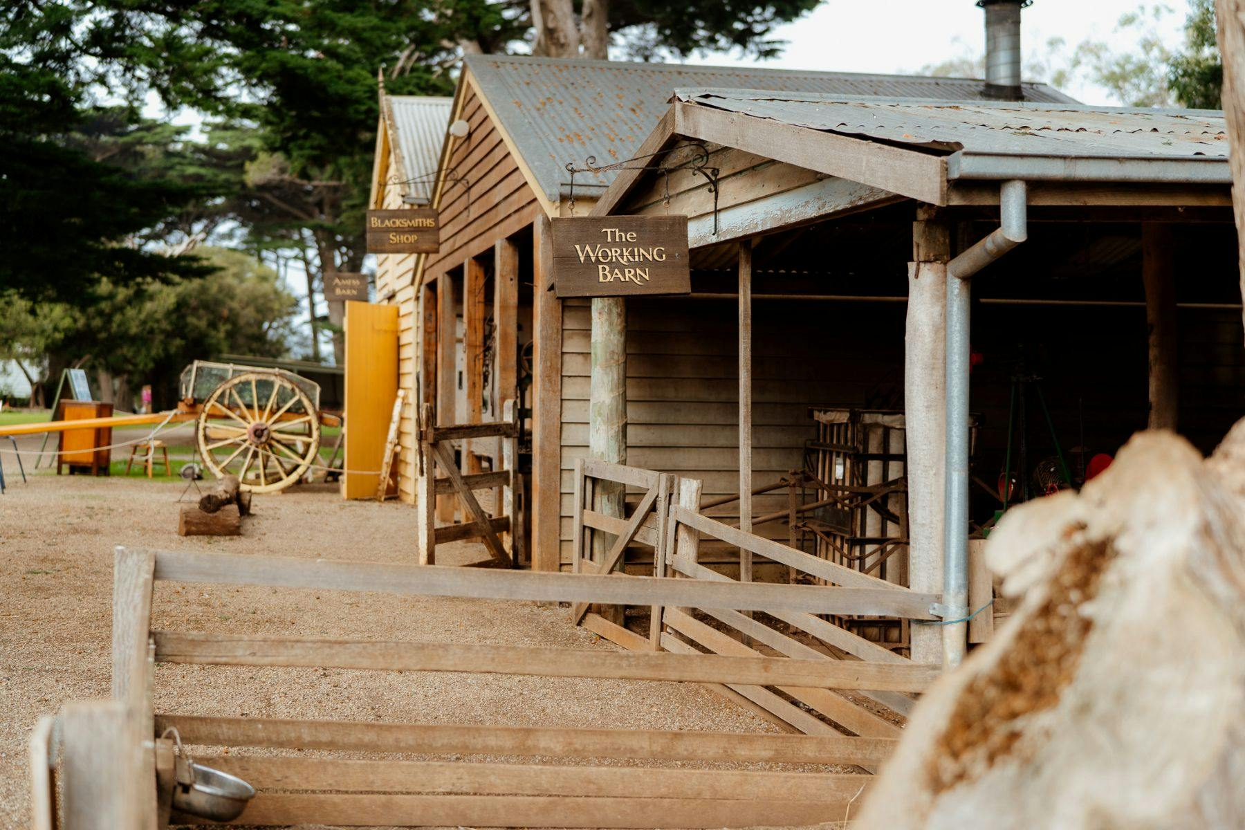 Grange en bois avec un panneau indiquant "The Working Barn" à côté de "Blacksmith's Shop". Une roue de chariot en bois et une clôture se trouvent à proximité.