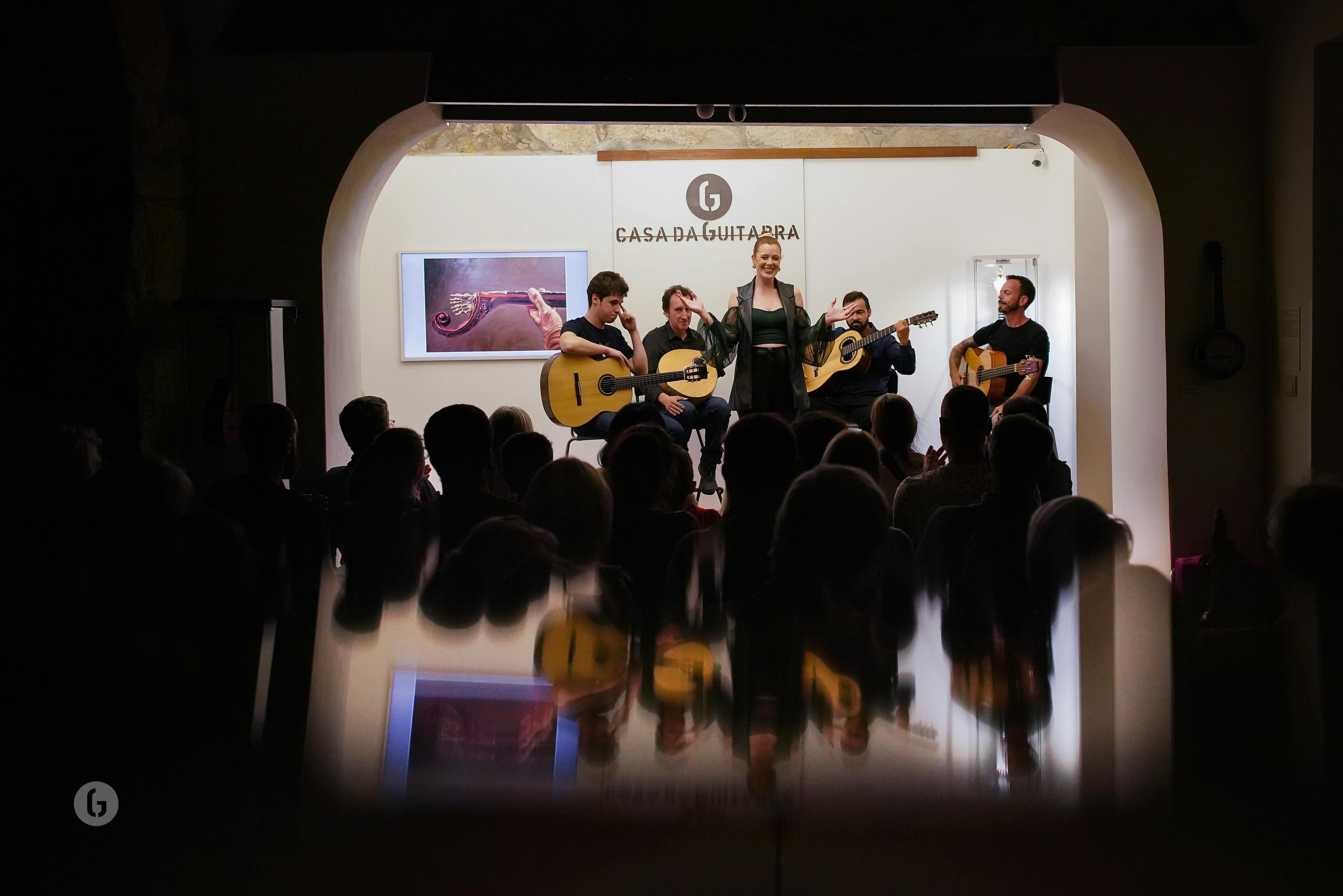 A musical performance with a woman singing and four guitarists on stage, watched by an audience at "Casa da Guitarra."