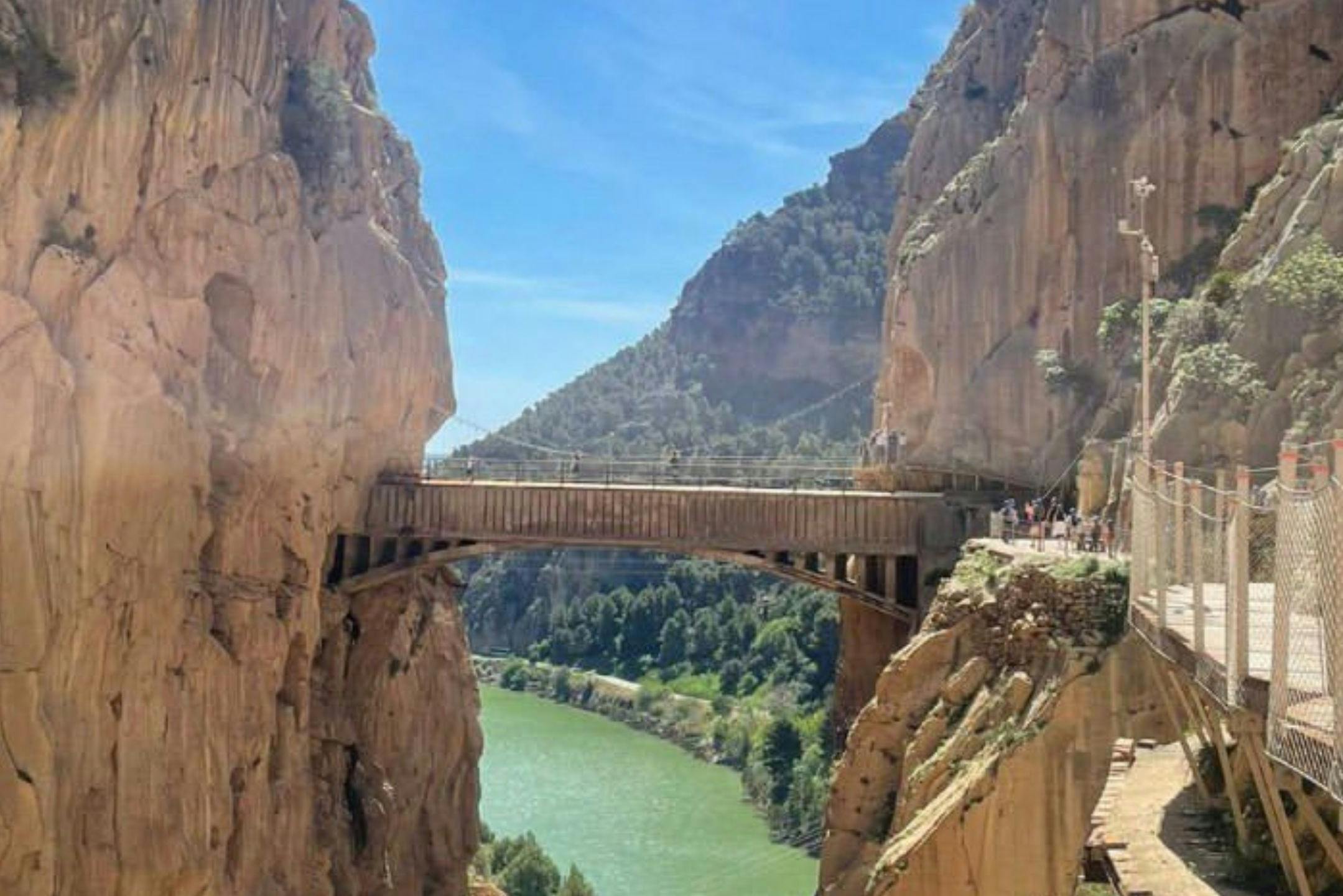 A scenic view of a bridge spanning a narrow canyon with a river below, surrounded by cliffs and a clear blue sky overhead.