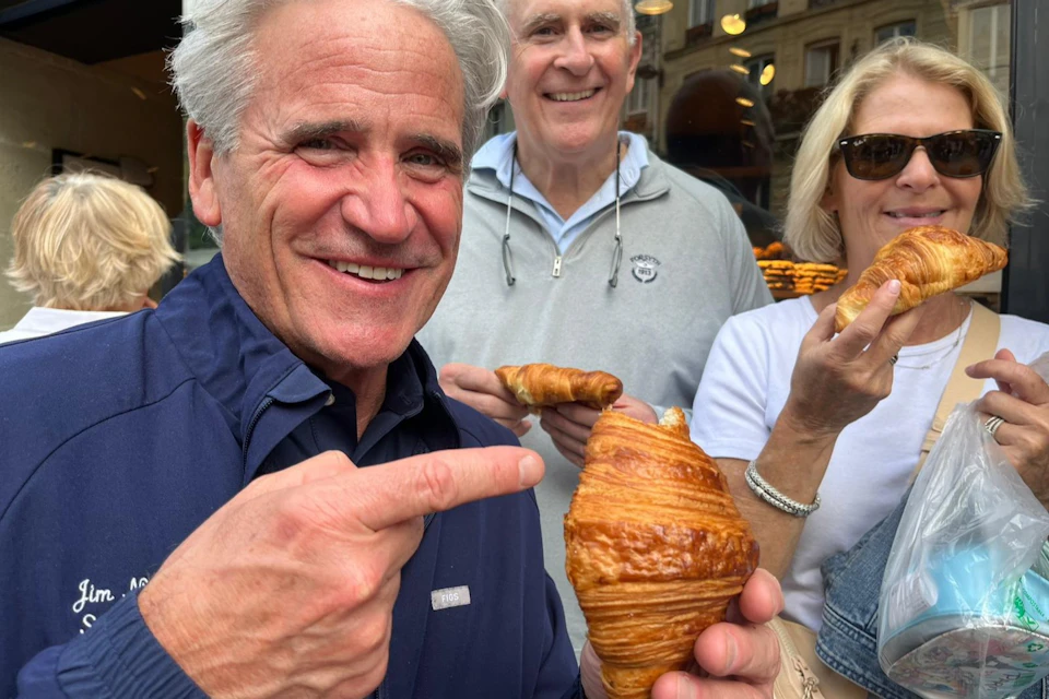 Three people smiling and holding croissants in front of a bakery.
