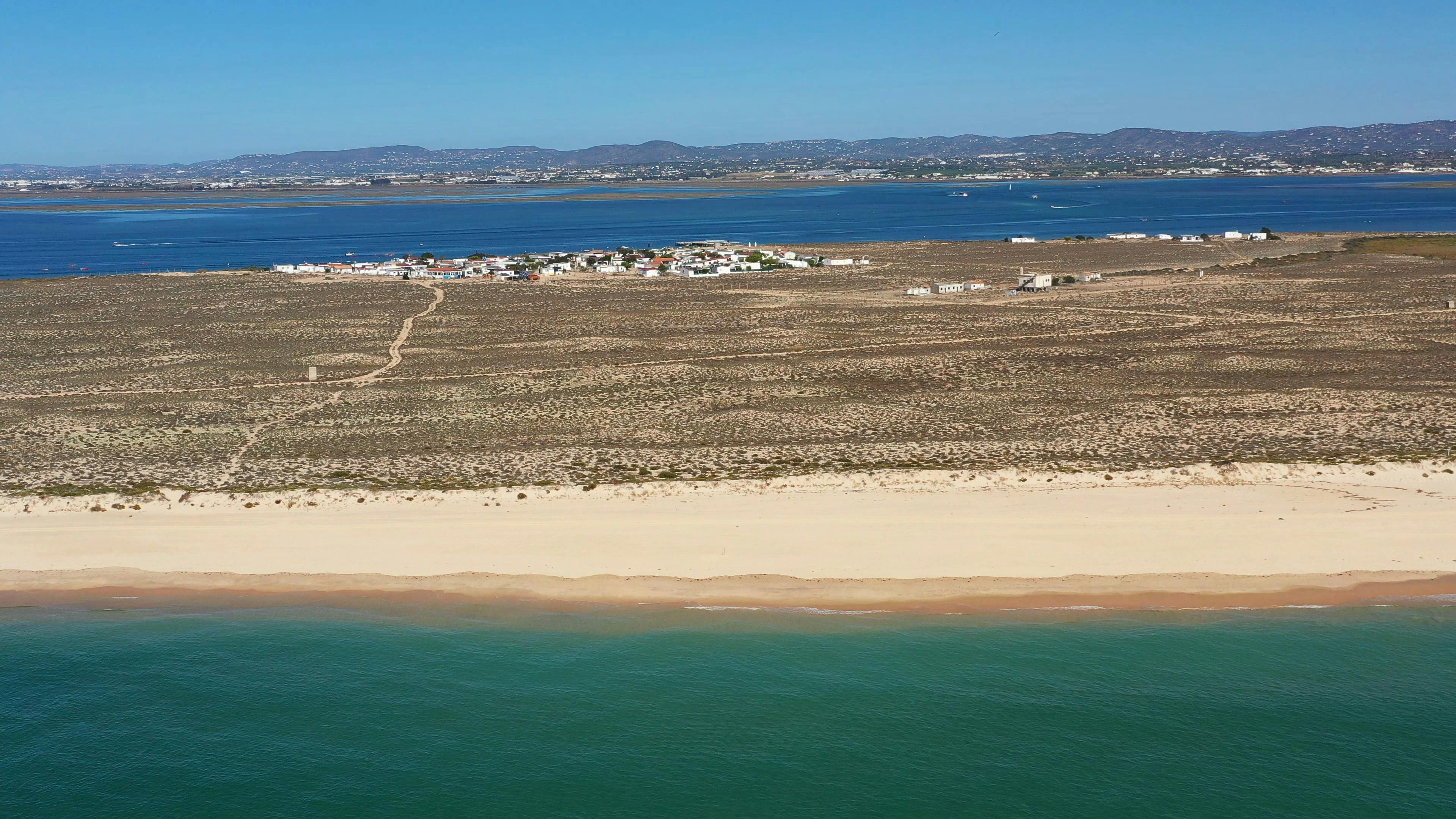 Αεροφωτογραφία αμμώδους παραλίας, με άγονο τοπίο και μικρό οικισμό σε απόσταση κοντά σε υδάτινο όγκο, κάτω από καθαρό ουρανό.