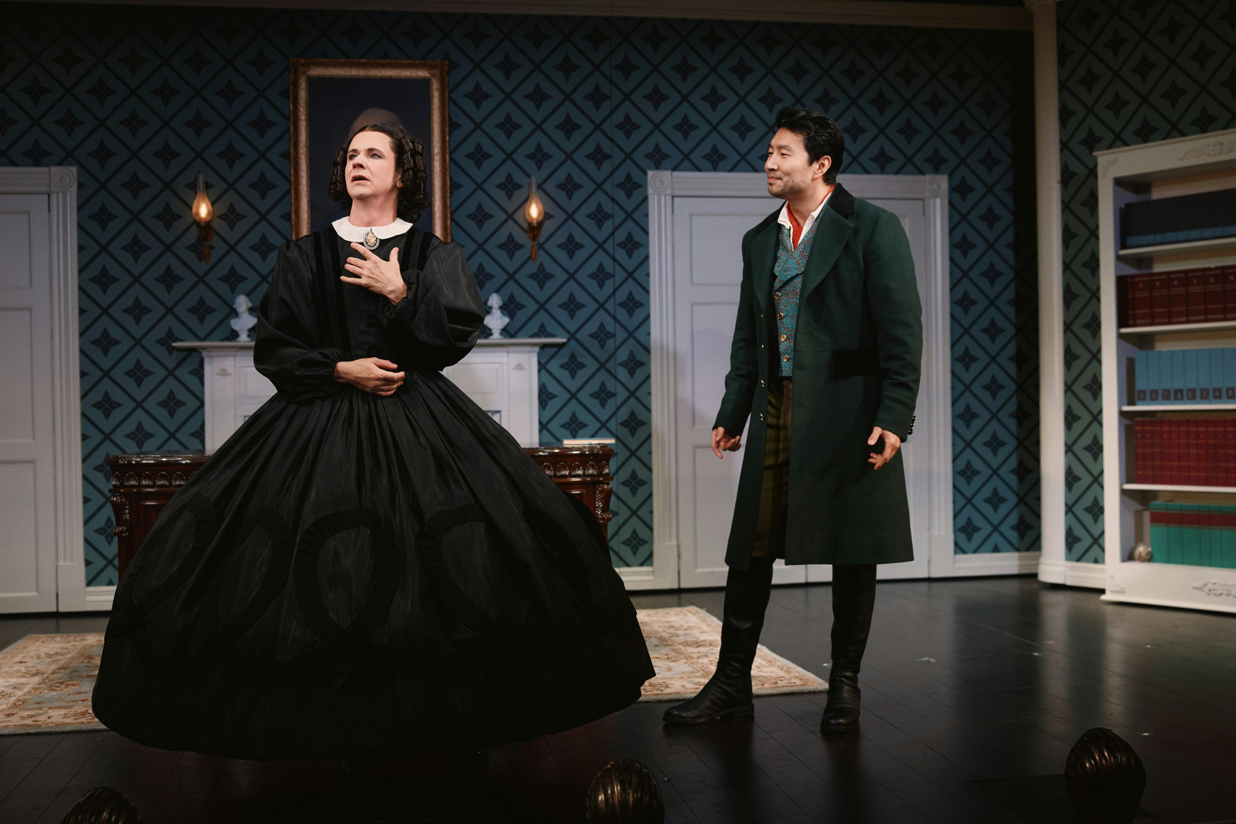 Two actors on a stage set resembling a vintage room, with patterned wallpaper, a bookshelf, and period costumes.