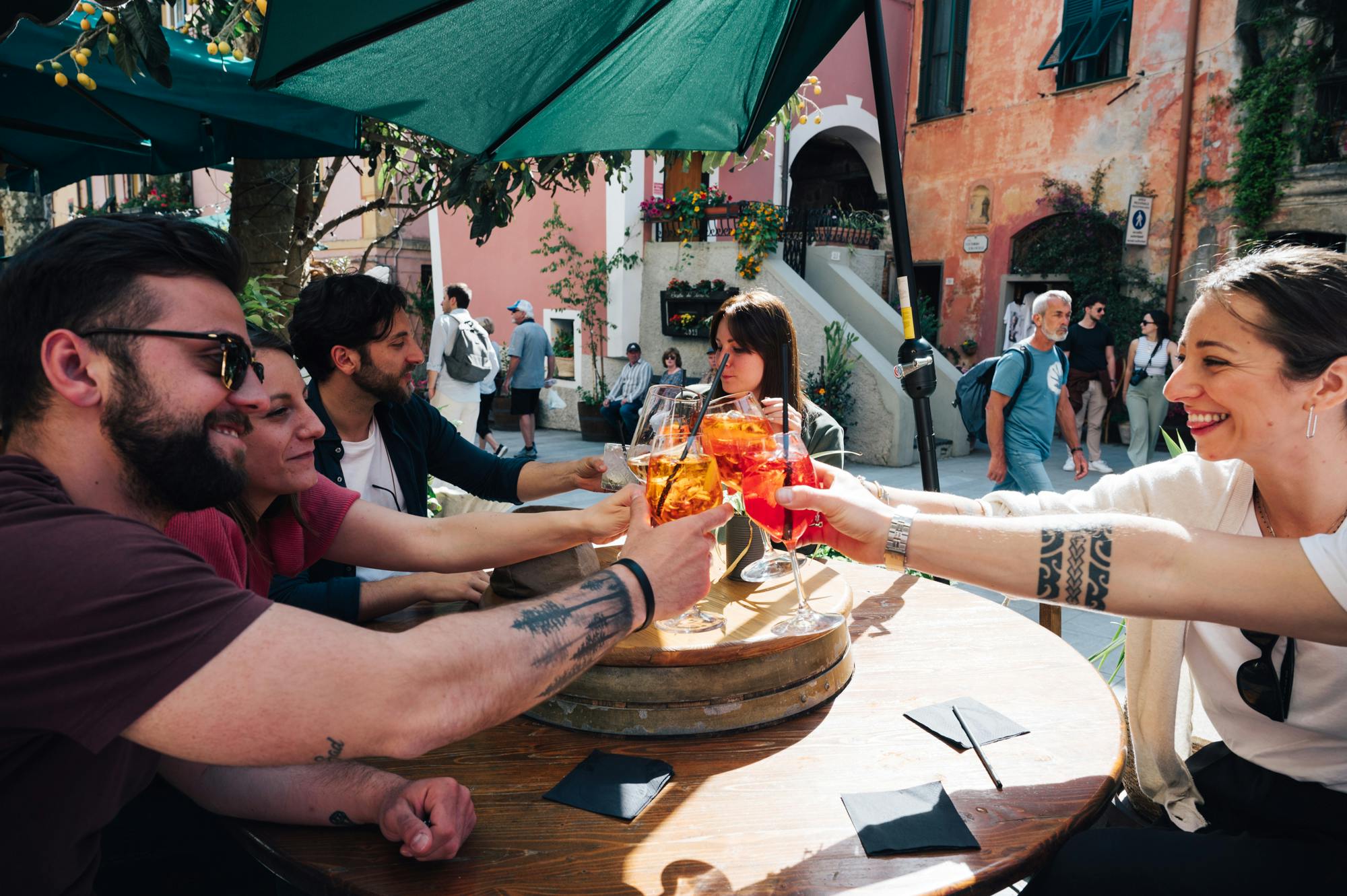 人们坐在户外咖啡馆，在阳光明媚的日子里举杯祝酒，背景是一栋质朴的建筑。